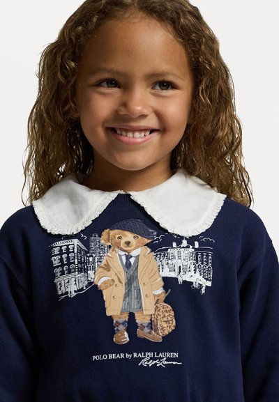 Mornarica jemnica s beelim nabranim ovratnikom, sa crtežem medveda u bež kaputu i odjeći, u pozadini su konture zgrada.