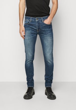 Mann trägt eng anliegende blaue Jeans mit ausgebleichten Oberschenkeln, schwarzes Hemd und schwarze Schnürstiefel, steht mit einer Hand in der Tasche.