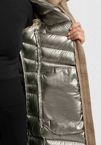 Doudoune légère avec un intérieur argenté métallisé et un extérieur beige. Présente une texture matelassée et une poche intérieure.