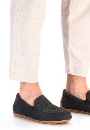 Black suede loafers with perforated design, flexible soles, and tan rubber outsoles. Worn with light-colored, plaid fabric pants.