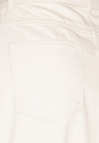 Valkoiset farkut, joissa on takatasku neliönmuotoisella läpällä ja hienovaraisilla ompeluyksityiskohdilla, esittelevät sileää, strukturoidun näköistä pintaa.