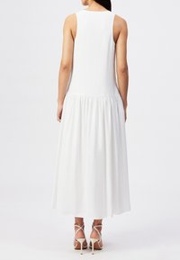 Kvinde iført en ærmeløs, hvid kjole til anklerne med en rynket nederdel og beige højhælede sandaler, vist bagfra.