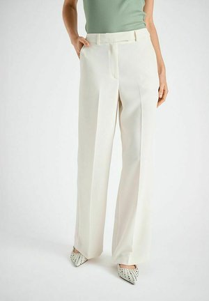 Personne portant un pantalon blanc taille haute à jambes larges, un haut sans manches vert pâle, et des chaussures blanches pointues à pois noirs, debout sur un sol blanc.