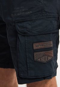Marineblå cargoshorts lavet af blødt stof, med synlige syninger, flere lommer og broderede lapper med tekst og mønstre.