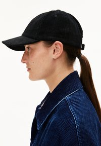 Schwarze Cordhüte mit einem gekrümmten Schirm, der eine strukturierte Oberfläche und einen verstellbaren Riemen hinten hat. Getragen mit einer blauen Cordjacke.