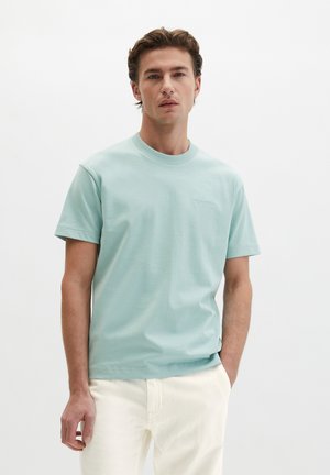 Jonge man met kort bruin haar, gekleed in een lichtblauw T-shirt met korte mouwen en ronde hals, en een witte broek, staand met één hand in zijn zak.