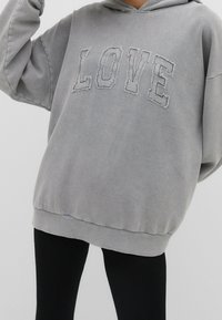 Felpa grigia oversize con tasca a marsupio e ricamo testurizzato "LOVE" sul fronte. Tessuto morbido e orlo a coste. Indossata con leggings neri.