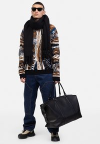 Strickpullover mit geometrischen Mustern in Braun, Blau und Creme; dunkle Denim-Hose; schwarzer Schal; schwarze Leder-Duffle-Bag; grau-schwarze Sneakers.