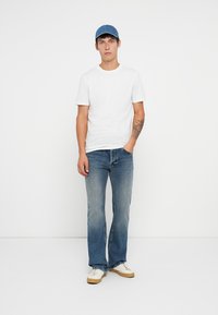 T-shirt branco de manga curta, jeans de ganga azul escuro com ligeira flare, boné azul e ténis brancos com detalhes em bege. Design simples.