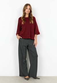 Pulover oversized burgundy cu mâneci largi, asortat cu pantaloni largi gri închis și tocuri alb-negru cu dungi. Materiale texturate vizibile.