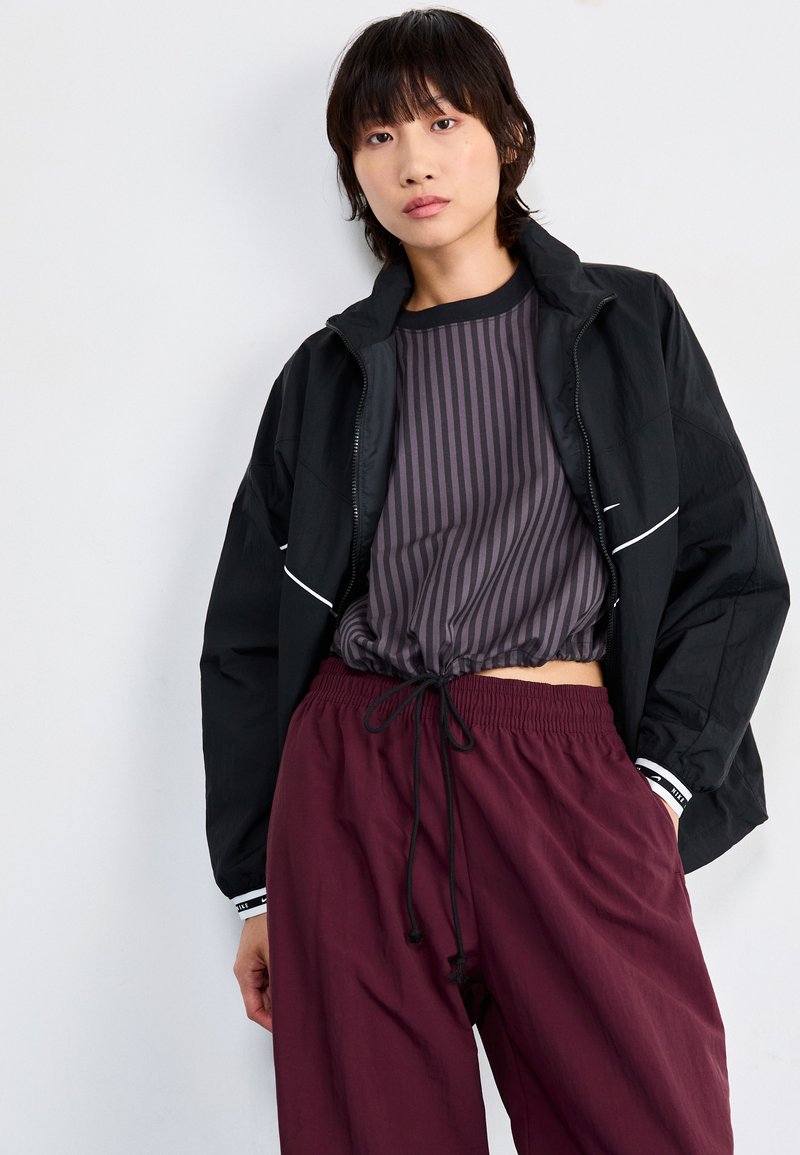 Giacca nera con zip, maglietta a righe viola con cordoncino e pantaloni elasticizzati bordeaux in vita. Caratteristiche includono dettagli bianchi e vestibilità comoda.