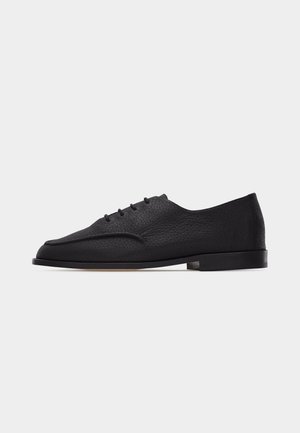 Chaussure Oxford en cuir texturé noir avec petit talon, bout arrondi et laçage fermé sur un fond blanc uni.