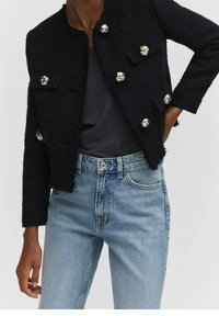 Giacca nera con texture, dotata di bottoni argentati e tasche, indossata sopra un top grigio e jeans azzurri chiari. Design corto e aderente con orli sfilacciati.