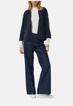 Vrouw draagt een witte coltrui, een marineblauw open jack met zakken aan de voorkant, wijd uitlopende donkerblauwe jeans en witte sneakers, staand tegen een effen achtergrond.