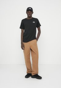 Μαύρο βαμβακερό μπλουζάκι με λευκό λογότυπο, χακί παντελόνι με φαρδιά πόδια και μαύρα αθλητικά παπούτσια, συμπληρωμένα με μαύρο καπέλο που ταιριάζει. Απλός και casual σχεδιασμός.