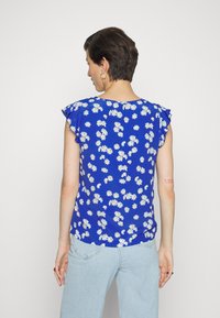 Top floral azul com padrões de margaridas brancas, mangas curtas e franzidas, e um acabamento curvo, combinado com jeans azul claro.