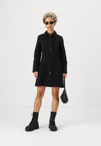 Crna traper haljina košulja s ovratnikom, kopčanjem sprijeda, dugim rukavima i prilagođenim krojem. Uparena s crnim čizmama na vezanje i malom torbicom.