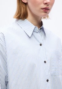 Γυναίκα που φοράει λευκό πουκάμισο με λεπτές κατακόρυφες μπλε ρίγες, κουμπωμένο μπροστά, με ριγέ τσέπη στο στήθος, κομμένο κάτω από τη μύτη.