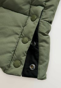 Olivenfarvet polstret jakke med store sorte trykknapper og sort inderfor; blød tekstur med kviltet design.