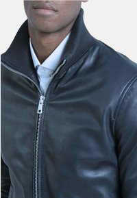 Zwarte leren motorjack met een hoge geribbelde kraag, zilveren rits en een gladde textuur, gedragen over een wit overhemd met kraag.