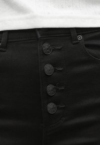 Črne jeans hlače z visokim pasom iz jeansa, z osem gumbi na sprednji strani, stranskimi žepi in gladko teksturo. Tkanina je rahlo obrabljena.