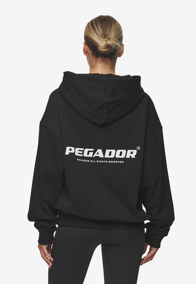 Sudadera negra de gran tamaño con capucha, que presenta un logo blanco impreso "PEGADOR" en la parte posterior. Puños y dobladillo acanalados. Textura de tela suave.