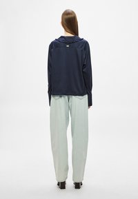 Μπλε ναυτικό αμάνικο πανωφόρι με βολάν στον γιακά, μακριά μανίκια και άνετη εφαρμογή, σε συνδυασμό με ανοιχτό πράσινο παντελόνι φαρδιάς γραμμής και μαύρα χοντρά τακούνια.