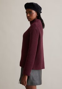 Burgunderfarbener Strickpullover mit hohem Kragen und strukturiertem Muster, kombiniert mit einem schwarzen Lederrock. Das Model trägt eine schwarze Baskenmütze.