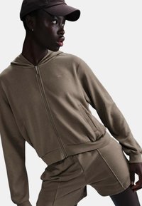 Felpa con zip marrone realizzata in materiale morbido, abbinata a pantaloni corti coordinati. Presenta un cappuccio, tasche laterali e un logo discreto sul petto.