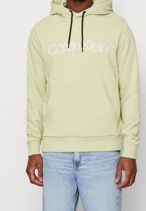 Osoba ubrana w jasnozieloną bluzę z kapturem Calvin Klein z czarnymi troczkami i jasnoniebieskie dżinsy, stojąca na jednolitym tle.