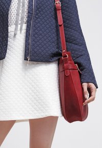 Borsa crossbody in pelle rossa con finitura testurizzata, caratterizzata da una forma arrotondata e una tracolla regolabile, abbinata a una giacca blu navy trapuntata e una gonna bianca.