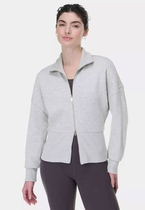 Vrouw met gevlochten haar die een lichtgrijze sweater met rits draagt over donkere aansluitende leggings, staand tegen een eenvoudige witte achtergrond.