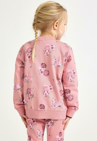 Młoda dziewczyna z blond warkoczami, ubrana w różową bluzę i spodnie w fioletowo-różowe wzory róż, stojąca tyłem.