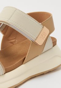 Beige og kremfargede plattformsandaler med teksturerte stropper og en gullfarget aksent. Mykt fotseng med subtil søm og gummisåledesign.
