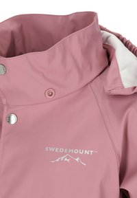 Veste imperméable rose avec un col haut, des pressions argentées et une doublure blanche. Présente un logo avec un design de montagne sur le devant.