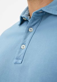 Lyseblå polo shirt i struktureret stof, med spidse kraver og tre knaplukninger. Bemærkelsesværdige detaljer inkluderer kontrasterende knapper.