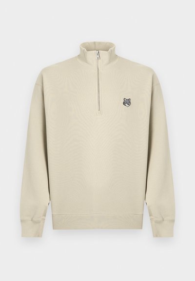 Μπεζ φούτερ με φερμουάρ στο ένα τέταρτο, μακριά μανίκια και ριμπ μανσέτες, με μικρό λογότυπο κεφαλής λύκου στο αριστερό μέρος του στήθους.