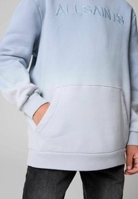 Ljusblå sweatshirt med gradientdesign, utrustad med en broderad logotyp, en känguruficka och ribbad mudd och nederkant. Mjuk textur.