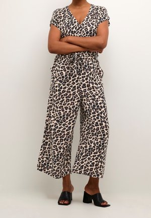 Kvinde stående med armene krydsede iført leopardmønstret buksedragt med korte ærmer og sorte hælede mule-sandaler med åben tå mod en ensfarvet baggrund.