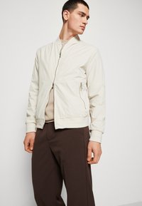 Giacca bomber beige chiaro con finitura texture, polsini e colletto a coste, chiusura con zip sul davanti e tasche laterali, abbinata a pantaloni marroni.