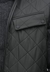 Czarna pikowana kamizelka z zamkiem błyskawicznym, naszywaną kieszenią na zatrzaski oraz widocznym pod spodem szarym swetrem o fakturze.