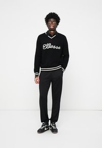 Sort strikket sweater med hvide striber og "Ellesse"-logo, parret med sorte bukser og sorte atletiksko. Enkel, sporty design.