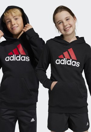 Sudaderas negras con un gran logo de Adidas en blanco y rojo, con un bolsillo frontal y puños de canalé. Llevadas por un niño y una niña.