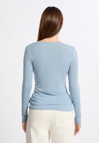 Maglietta a maniche lunghe di colore azzurro chiaro realizzata in tessuto morbido, con scollatura a barchetta e dettaglio arricciato sul retro, indossata con pantaloni color crema.