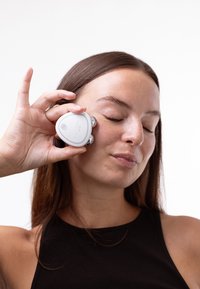 Weißer Gesichtsmasierer mit Metallakzenten, ovale Form, wird mit der Hand gegen die Wange geführt. Benutzer hat die Augen geschlossen und eine helle Haut.