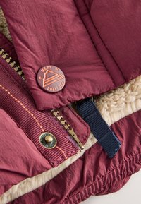 Section d'une doudoune bordeaux avec une doublure en polaire texturée. Comprend une fermeture éclair dorée, des coutures décoratives orange et un bouton accessoire de marque.