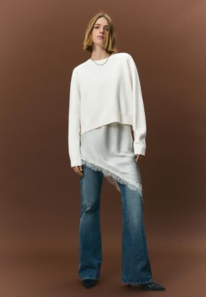 Biela krátka mikina nosená cez bledý saténový asymetrický top so čipkovaným lemom, doplnená rozšírenými modrými džínsami a špicatými čiernymi lodičkami.