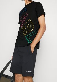 Osoba v černém tričku Karl Lagerfeld se barevnými písmeny a černých šortkách Dondup, držící černou a zelenou tašku.