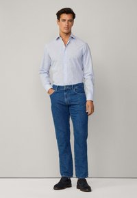 Camisa de botones a rayas azul claro y blanca con mangas largas, combinada con jeans de mezclilla azul oscuro y zapatos oscuros. Diseño simple, ajuste entallado.