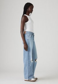 Jean large bleu clair à l'aspect usé avec des ourlets effilochés et des déchirures visibles, associé à un débardeur blanc et des chaussures de sport marron.